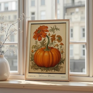 Op de afbeelding: Een ingelijste kunstafdruk met een levendige oranje pompoen en bloemen, met de tekst "William Morris" en "Pumpkin". Het kunstwerk staat tegen een crèmekleurige achtergrond, is omlijst in licht hout en wordt op een vensterbank getoond.
