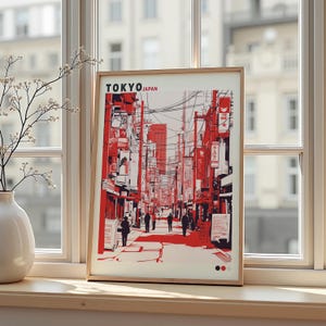 Może przedstawiać: Oprawiony wydruk przedstawiający czerwoną i białą ilustrację sceny ulicznej w Tokio. Wydruk zawiera tekst "TOKYO JAPAN" na górze. Grafika jest eksponowana na parapecie, obok białego wazonu z kompozycją kwiatową.