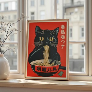 May include: A framed art print featuring a black cat eating noodles from a bowl. The cat has yellow eyes and is set against a red background. The bowl and background have Japanese text. The print is displayed on a windowsill.