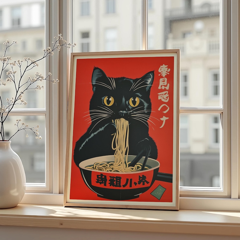 May include: A framed art print featuring a black cat eating noodles from a bowl. The cat has yellow eyes and is set against a red background. The bowl and background have Japanese text. The print is displayed on a windowsill.