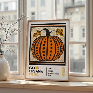 Op de afbeelding: Een ingelijste kunstdruk met een oranje pompoen met zwarte stippen en een zwarte steel. Het kunstwerk bevat de tekst "YAYOI KUSAMA" en "JAPAN 1997 PUMPKIN SERIES No.2". De print wordt op een vensterbank getoond.