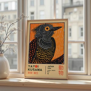 Op de afbeelding: Een ingelijste kunstprint met een gestileerde vogelillustratie in zwart, oranje en geel. Het kunstwerk bevat de tekst "YAYOI KUSAMA" en Japanse karakters. De vogel zit op een tak tegen een oranje achtergrond.