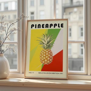 Könnte beinhalten: Ein gerahmter Kunstdruck mit einer Ananas-Illustration. Das Wort "PINEAPPLE" steht oben in fetten schwarzen Buchstaben. Das Bild hat einen geometrischen Hintergrund in Gelb, Grün und Rot. Der Text "BAUHAUS EXHIBITION 1951" steht unten.
