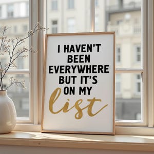 Może przedstawiać: Oprawiony wydruk z tekstem "I HAVEN'T BEEN EVERYWHERE BUT IT'S ON MY list". Tekst jest czarny i złoty na białym tle. Oprawiony wydruk jest wyświetlany na parapecie.