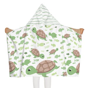 May include: A white hooded towel with a pattern of green turtles and fish. The turtles have brown shells and green bodies. The hood has green stripes. The towel is designed for children.