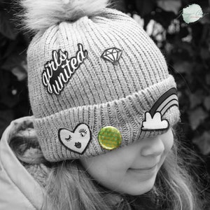 Op de afbeelding: Een grijze gebreide beanie met een pompon bovenop. De beanie heeft verschillende patches erop, waaronder een hart met een knipperend oog, een regenboog, een diamant en een patch met de tekst "girls united".