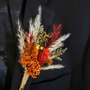 May include: A colorful boutonniere featuring dried flowers in shades of orange, red, and cream, accented with small yellow blooms. The stems are wrapped with twine, and the arrangement is pinned to a black suit jacket. A rustic, natural accessory.