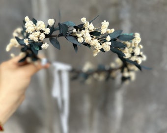 May include: A white flower crown with green leaves. The crown is made of dried flowers and is perfect for a wedding or other special occasion.