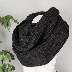 以下が含まれることがあります： マネキンに展示された、黒色のニットインフィニティスカーフ。スカーフは厚手で、リブ編みの模様があります。マネキンは白いトップスと黒いショートパンツを着用しています。背景は、鉢植えの植物がある白い壁です。