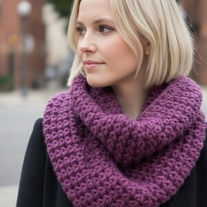以下が含まれることがあります： 紫色のかぎ針編みインフィニティスカーフのクローズアップ。スカーフは深い紫の色合いで、太い糸で作られているようです。かぎ針編みの質感が確認でき、スカーフは首にかけられています。