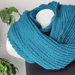 Op de afbeelding: Een tealblauwe gebreide oneindige sjaal. De sjaal is gemaakt met een eenvoudig steekpatroon en is zacht en comfortabel.