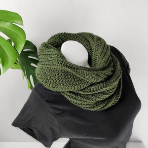以下が含まれることがあります： 濃い緑色のクロシェのインフィニティスカーフ。スカーフは厚手で質感があり、半分に折り畳んでループを作っています。