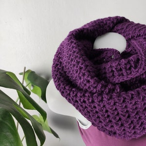 以下が含まれることがあります： 紫色のクロシェの無限スカーフ。スカーフは太い糸で作られており、質感のあるステッチパターンがあります。