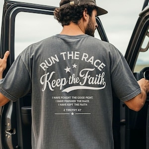 May include: A charcoal gray short-sleeve t-shirt featuring white text on the back that reads RUN THE RACE Keep the Faith, followed by the scripture I HAVE FOUGHT THE GOOD FIGHT. I HAVE FINISHED THE RACE. I HAVE KEPT THE FAITH 2 TIMOTHY 4:7.