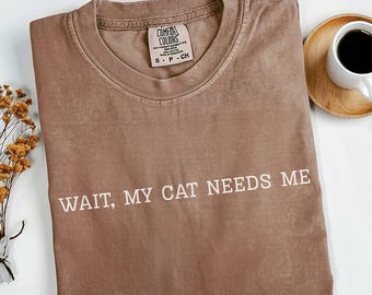 May include: Espresso-colored t-shirt with the text "WAIT, MY CAT NEEDS ME" in white. The shirt is folded and has a Comfort Colors tag. A cup of coffee and a small bouquet of dried flowers are in the background.