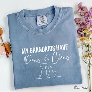 May include: A blue crew neck t-shirt featuring the white text MY GRANDKIDS HAVE Paws & Claws above a simple line art illustration of a dog and a cat. The shirt has a Comfort Colors brand label on the inside collar and a short-sleeved design.