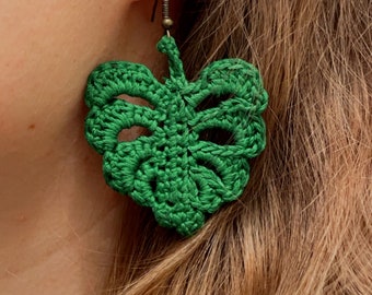 May include: A pair of green crocheted earrings shaped like monstera leaves. The earrings are hanging from the earlobes of a person with long brown hair.