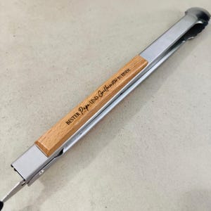 Könnte beinhalten: Grillzange aus Edelstahl mit Holzgriff. Der Griff trägt die Gravur "BESTER Papa UND Grillmeister IN EINEM". Die Zange ist für das Grillen und Kochen im Freien konzipiert.