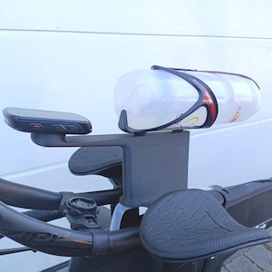 May include: A black and gray bicycle handlebar setup with a water bottle, armrests, and a small electronic device. The water bottle is clear with red and black accents. The armrests have a textured surface.