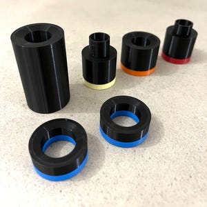 Puede incluir: Una colección de objetos cilíndricos negros impresos en 3D con varios acentos de color. Algunos tienen bases o anillos azules, naranjas, rojos o crema. Los objetos varían en tamaño y forma, con interiores roscados.