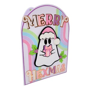 May include: A pastel pink and purple card with a rainbow design. The card features a ghost wearing a Santa hat and holding a gift. The text "Merry Hexmas" is written on the card.