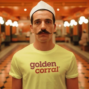 May include: A man wearing a yellow t-shirt with the words "golden corral" in burgundy. He has a white hat and a large mustache. The background is an orange-toned restaurant.