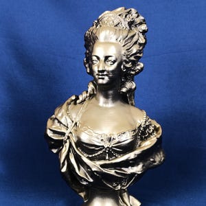 Peut inclure: Buste doré d'une femme avec une coiffure élaborée et des vêtements d'époque. La sculpture présente des traits de visage détaillés, une écharpe drapée et une base de piédestal. Le buste est placé sur un fond bleu uni.