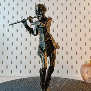 Può includere: Una scultura in metallo dorato di una persona che suona il flauto. La scultura è realizzata con forme geometriche e ha una superficie testurizzata.