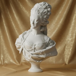 May include: A white bust sculpture of a woman with an elaborate hairstyle, adorned with roses and pearls. The sculpture features detailed clothing and is mounted on a pedestal. The background is a gold-colored fabric.