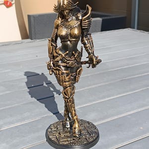 Può includere: Statuetta color bronzo di una guerriera in armatura, con una spada. La figura è su una base circolare con un motivo a ciottoli. L'armatura presenta dettagli intricati e la figura ha capelli fluenti.
