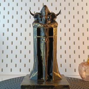 Peut inclure: Une statue en 3D imprimée en noir et or d'une figure tenant une épée. La figure porte une robe et a des cornes. La statue est sur une base noire et or.