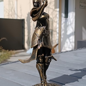 Può includere: Statuetta color bronzo di una donna in posa dinamica, con un braccio alzato. La figura è dettagliata con abiti fluenti e armature intricate. La statua è montata su una base circolare, mostrando arte e artigianato.