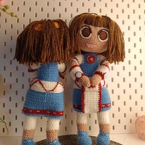 May include: Two crocheted dolls with brown hair, blue and white dresses, and red accents. The dolls are standing on a woven mat.