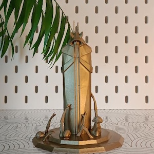 Peut inclure: Une sculpture géométrique dorée avec un corps rectangulaire et un sommet en forme de couronne. Plusieurs figures abstraites entourent la base. Des feuilles vertes sont dans le coin supérieur gauche. La sculpture est sur une surface à motifs.