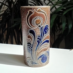 May include: A cylindrical ceramic vase featuring a textured brown and beige speckled finish. The surface is decorated with an embossed floral motif in deep blue and tan tones, characterised by swirling leaf patterns and a central stylised flower.