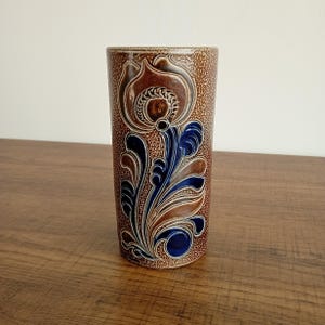 May include: A tall, cylindrical brown ceramic vase with a floral design in relief. The design features a large flower and stylized leaves in shades of brown, blue, and white. The vase has a textured surface and sits on a wooden table.