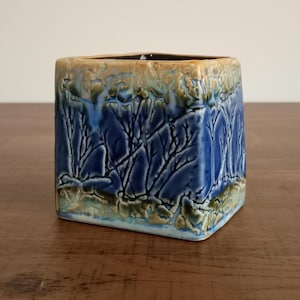 May include: Square ceramic vase with a textured, glazed finish. The vase has a blue and brown colour scheme with a tree-like pattern. The interior is dark, and the vase sits on a wooden surface.