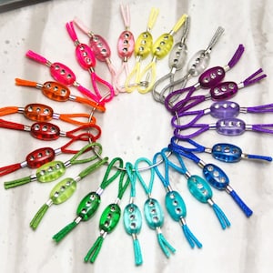 Puede incluir: Una colección de tiradores de cremallera coloridos con cuentas de plástico transparente y diamantes de imitación. Los tiradores de cremallera están dispuestos en un círculo sobre una superficie blanca.