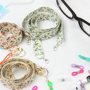 Może przedstawiać: Trzy sznurki z tkaniny z kwiatowym wzorem i srebrnymi klamrami, niebieska maska chirurgiczna, różowo-biała tkanina w kratkę, pomarańczowy sznurek z koralikami i numerami, czarne okulary do czytania, czarne okulary przeciwsłoneczne i różne kolorowe silikonowe uchwyty do okularów.