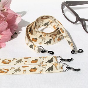 Puede incluir: Dos cadenas de gafas blancas con un diseño de estampado de gato. Las cadenas están hechas de tela y tienen cierres metálicos. Las cadenas están sobre una superficie blanca. También hay dos pares de soportes para gafas negros.