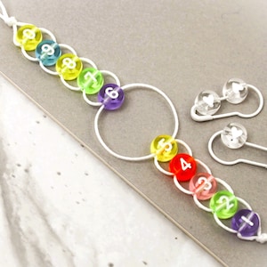 May include: A rainbow-colored beaded bracelet with a white string and a white circle charm. The beads are numbered 1 through 9. There are also two safety pins with clear beads and white hearts.