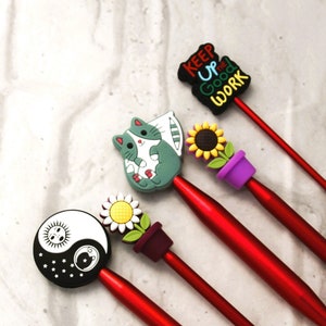 以下が含まれることがあります： 異なる色のプラスチック製の先端を持つ赤い編み針が5本。先端には、猫、ひまわり、陰陽のシンボル、デイジー、そして「Keep Up the Good Work」と書かれた看板が描かれています。