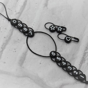 May include: Black cord with a loop at one end and numbered black beads. The cord has a circle shape in the middle. There are three separate black safety pins with white star and heart designs.
