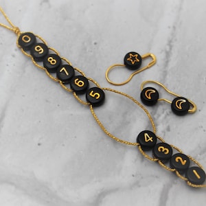 以下が含まれることがあります： 編み物用のアクセサリーセット。ステッチマーカーと段数カウンターが含まれています。ステッチマーカーは、星と三日月が特徴の黒いビーズが付いた金色の安全ピンです。段数カウンターは、金色の数字が付いた黒いビーズです。