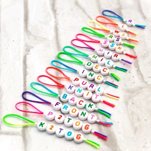 May include: A set of 15 colorful string bracelets with white letter beads spelling out knitting terms like "purl", "knit", "back", "front", "tog", and "k2tog".