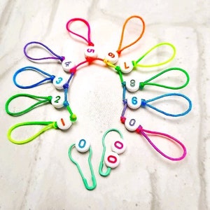 Könnte beinhalten: Ein Satz bunter Plastik-Maschenmarkierer mit den Zahlen 0-9 und dem Buchstaben L. Die Markierer sind an Schlaufen aus buntem Garn befestigt.