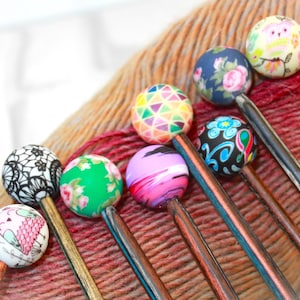 May include: A set of eight colourful knitting needles with decorative wooden handles and colourful, patterned beads. The beads feature various designs, including geometric patterns, floral motifs, and whimsical illustrations.