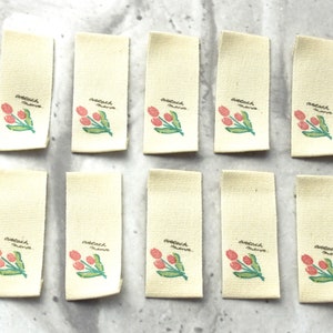 May include: Ten blank white fabric labels with a pink and green tulip design and the text "attach here" printed on each label.