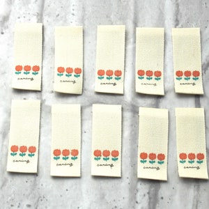 May include: Ten off-white fabric labels with a row of three orange flowers and green stems. Each label has the word "caring" written in black cursive.
