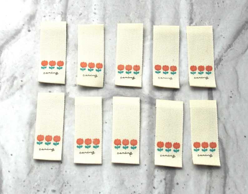 May include: Ten off-white fabric labels with a row of three orange flowers and green stems. Each label has the word "caring" written in black cursive.